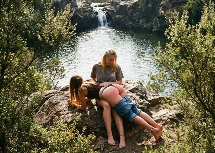 Spanking am Wasserfall: Junge braunhaarige Frau von blonder Frau draußen übers Knie gelegt